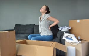 Woman (age 30-35) suffers from back pain due to unpacking boxes during a move into a new home. Moving house concept. Real people. copy space